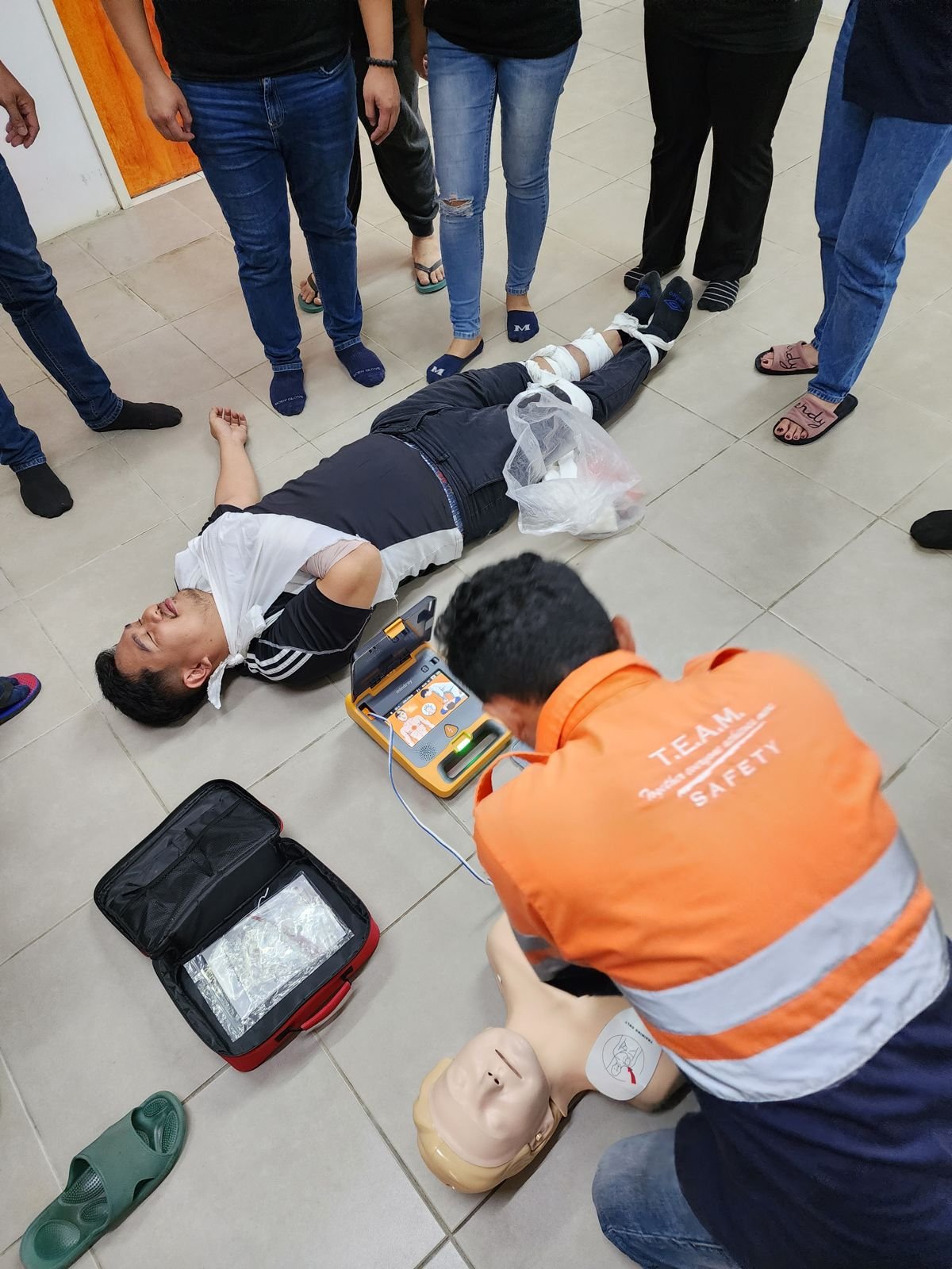 Overhead view of a trainer in an orange vest running a timed CPR and AED drill with a manikin and AED trainer